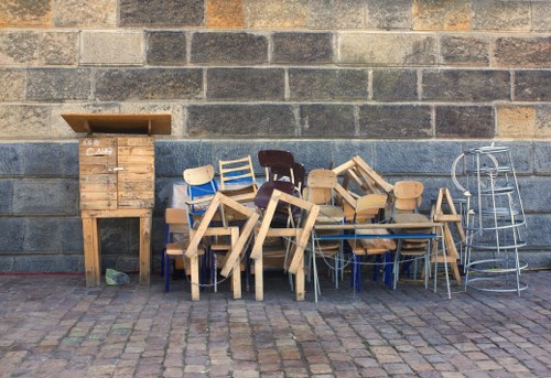 A collection of various discarded furniture and wooden items arranged on a paved area outdoors in front of a brick wall. The pile includes chairs with different designs and colors, such as light wood, dark wood, and painted surfaces, some stacked or leaning against each other. There are also wooden tables and small chests, one of which has a slanted top. To the right of the furniture, a metal collapsible drying rack or laundry rack is visible, standing upright. The scene appears to be part of a rubbish removal or clearance effort, possibly involving local services like those offered by Office Clearance Bow, situated near Bow or Stratford in east London, with the items awaiting disposal or recycling. The overall environment suggests an outdoor space adjacent to an urban or residential area, with the furniture and wooden items stacked along the brick wall on a cobbled surface, under natural daylight.