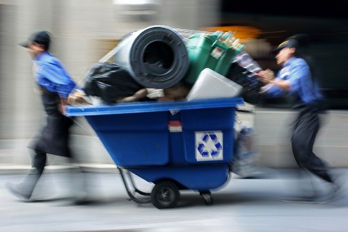 A blue waste disposal bin on wheels is shown being pushed along a city street by two individuals dressed in blue uniforms and black caps. The bin is overflowing with various rubbish items, including a large black trash bag, a green plastic storage container, a rolled-up carpet or mat, and other miscellaneous waste materials. The background features a blurred urban scene with indistinct buildings and vehicle movement, indicating motion and busy environment, likely in a town or city centre. The scene depicts a professional rubbish removal service, with the bin positioned on a paved sidewalk or kerbside area, emphasizing urban waste collection activities. The overall setting suggests an active effort to clear waste from commercial or residential premises, aligning with services provided by companies like Office Clearance Bow, operating within the Bow area, possibly around postcode E3 or nearby districts.