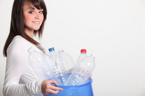 A young woman with shoulder-length dark brown hair and a light complexion is standing against a plain white background, smiling softly at the camera. She is wearing a white long-sleeved top and is holding a small blue plastic recycling bin filled with several clear plastic bottles, some with blue or red caps. The bottles are empty and vary in size, with some appearing partially crushed. The scene suggests an eco-friendly or rubbish collection context, emphasizing plastic waste disposal, which aligns with services offered by companies like Office Clearance Bow in the local area. The lighting is bright and even, highlighting the clear transparency and differing textures of the bottles, as well as the woman's relaxed posture and neat appearance, all contributing to a professional and straightforward depiction of waste management and recycling efforts.