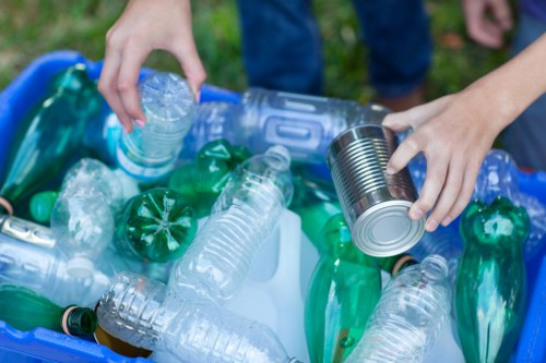 A person wearing blue jeans is seen placing a clear plastic bottle into a blue recycling bin filled with various other plastic bottles, including green and transparent ones, on a grassy outdoor area. The focus is on the hands handling the bottles, with the background showing a blurred patch of green grass and possibly other people nearby. The plastic bottles have textured surfaces and are of different sizes, some with caps and some without. The scene highlights waste sorting, aligned with rubbish removal services operating in local areas such as Bow, with the blue bin ready for collection. The natural daylight illuminates the scene evenly, emphasizing the clarity of the plastic bottles and the outdoor environment typical of a domestic or community recycling setting in London Borough of Tower Hamlets or nearby postcode areas.