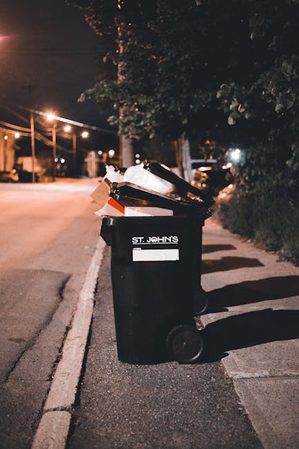 A collection of black plastic garbage bags filled with rubbish, positioned on a sidewalk near a black metal fence and a tree with dense foliage. The bags appear tightly packed and are placed close tog