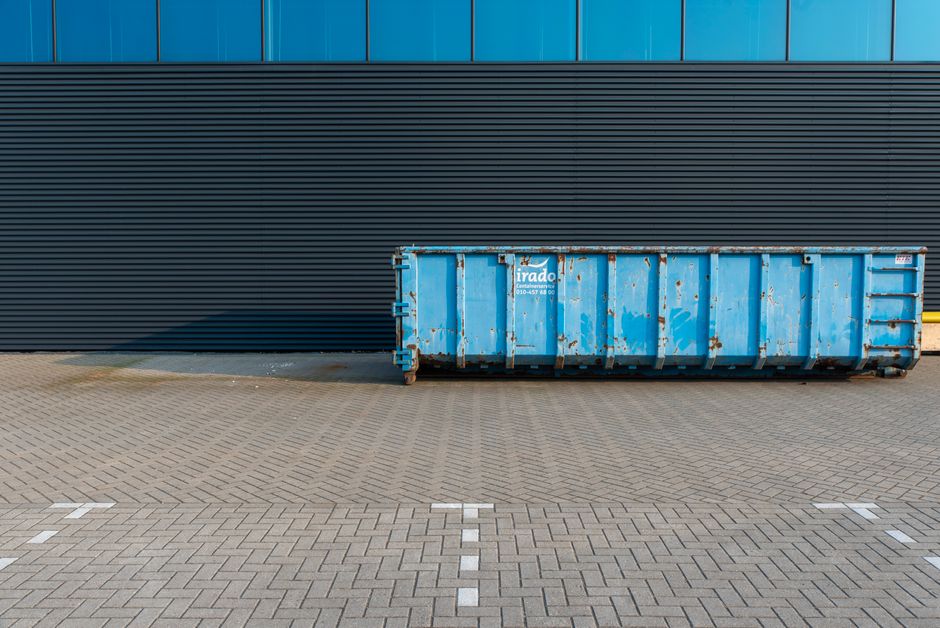 A row of large orange skip containers with green stripes and white labels on a paved outdoor area, positioned against a backdrop of leafless trees and a partly cloudy sky. The skips are made of metal,