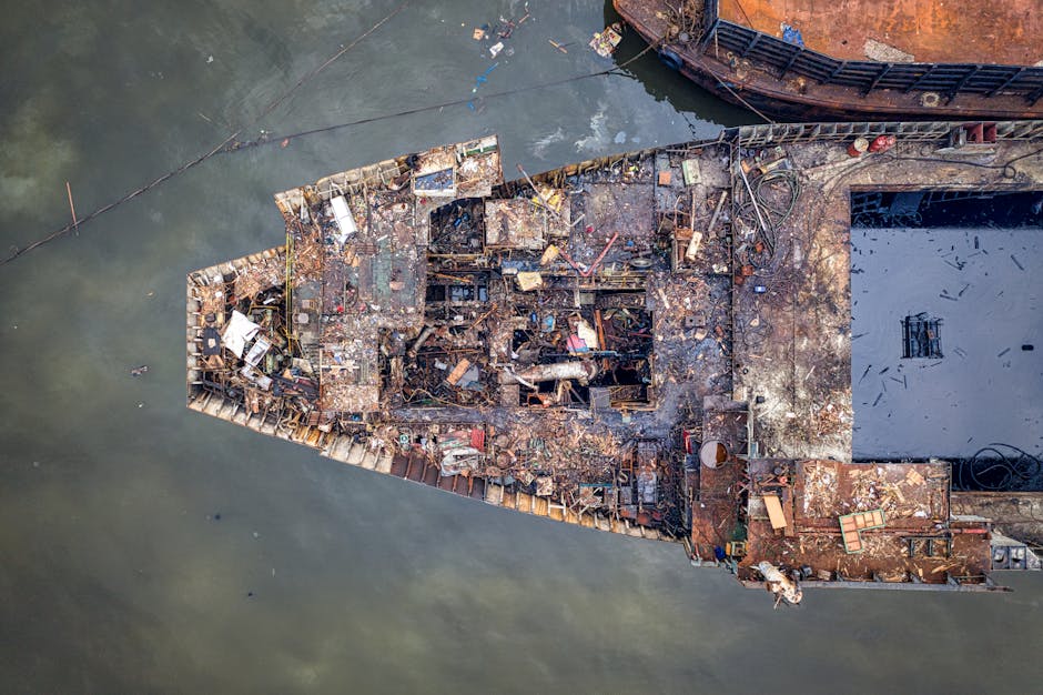 A group of workers employing manual tools and safety helmets are seen along the side of a large, rusted ship that is currently out of the water and positioned on land for repairs or dismantling. The s