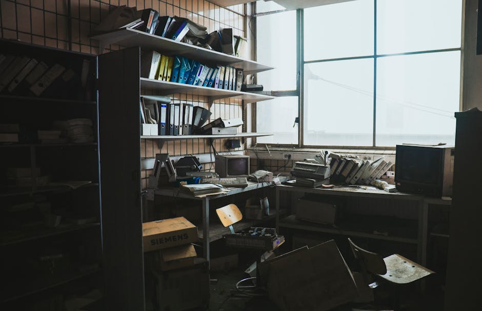The image depicts a modern office interior with several white partition panels separating individual workspaces. Each workspace contains a black office chair with a mesh backrest, a light-colored wood