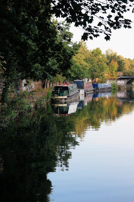 A narrow canal in an urban residential area during daytime, with several boats moored along the water's edge. The boats vary in size and color, with some showing visible weathered textures and metal o