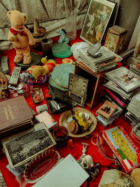 A cluttered indoor scene featuring various secondhand objects scattered across a red tablecloth. Visible items include a plush teddy bear wearing a red ribbon, several toy dinosaurs, a yellow rubber d